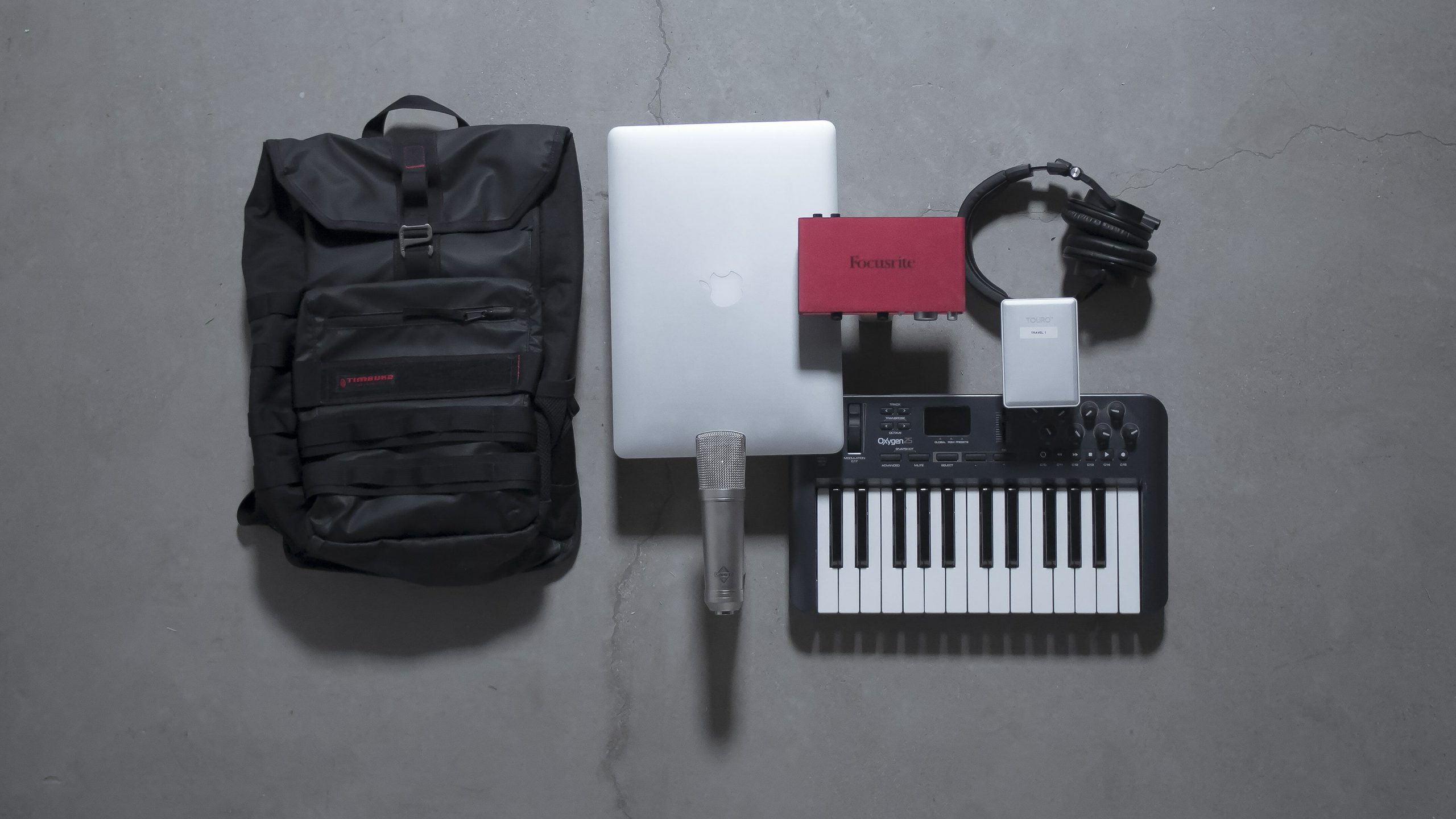 Travel essentials for musicians including a backpack, microphone, laptop, audio interface, headphones, portable keyboard, and portable hard drive